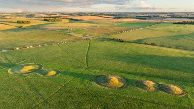 A wide aerial shot of the Stonehenge Landscape with The Cursus Barrows in the foreground and the monument visible further away in the distance.